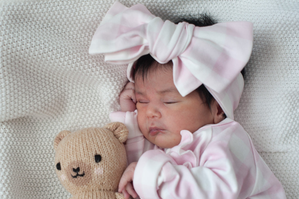 Baby in pink outfit with large bow headband holding a teddy bear on a soft surface Sweet June