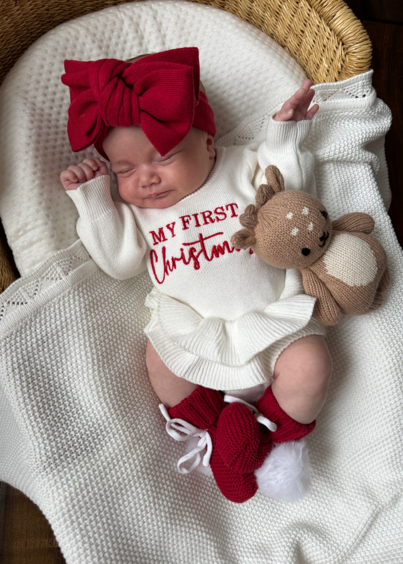 A sleeping newborn baby wearing a white knit romper with red text reading 'MY FIRST Christmas,' a large red bow headband, and red knit booties from Sweet June. 