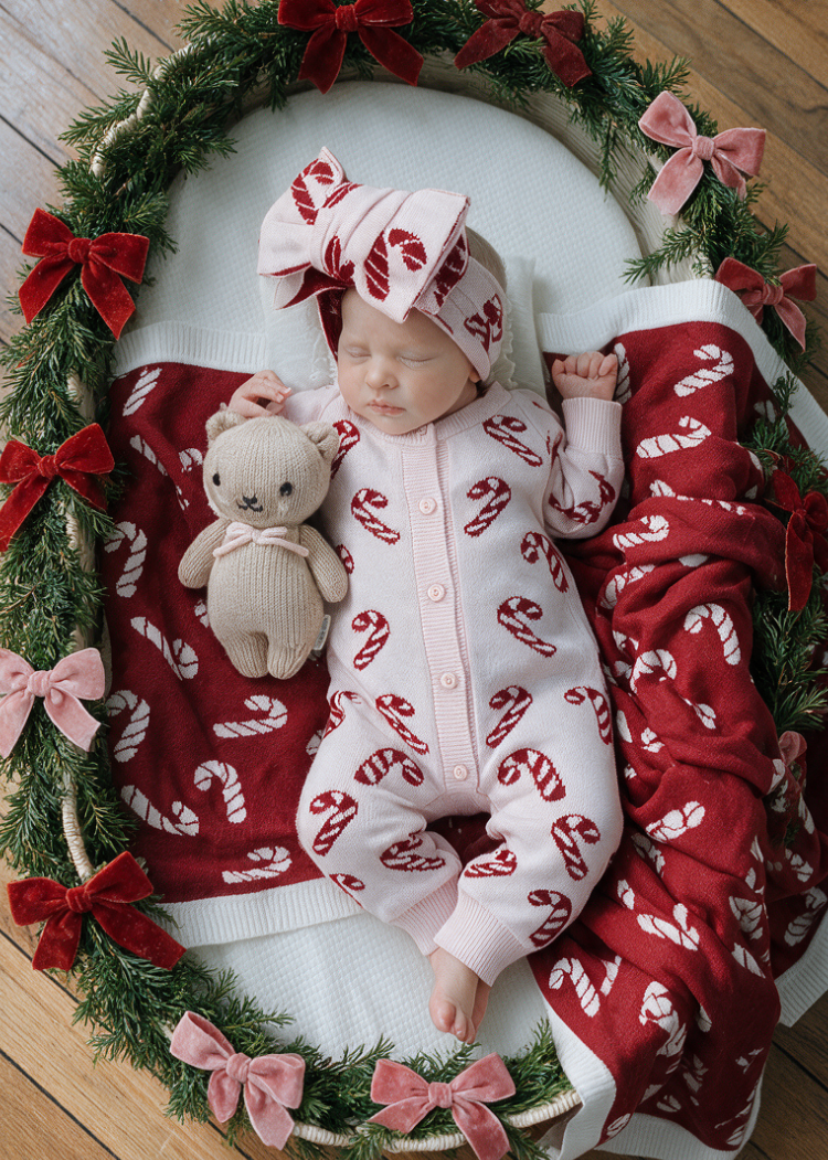 A newborn baby wearing a pink candy cane print footed sleeper and matching hat, lying on a white blanket inside a decorated wreath with red bows, next to a teddy bear from sweet june