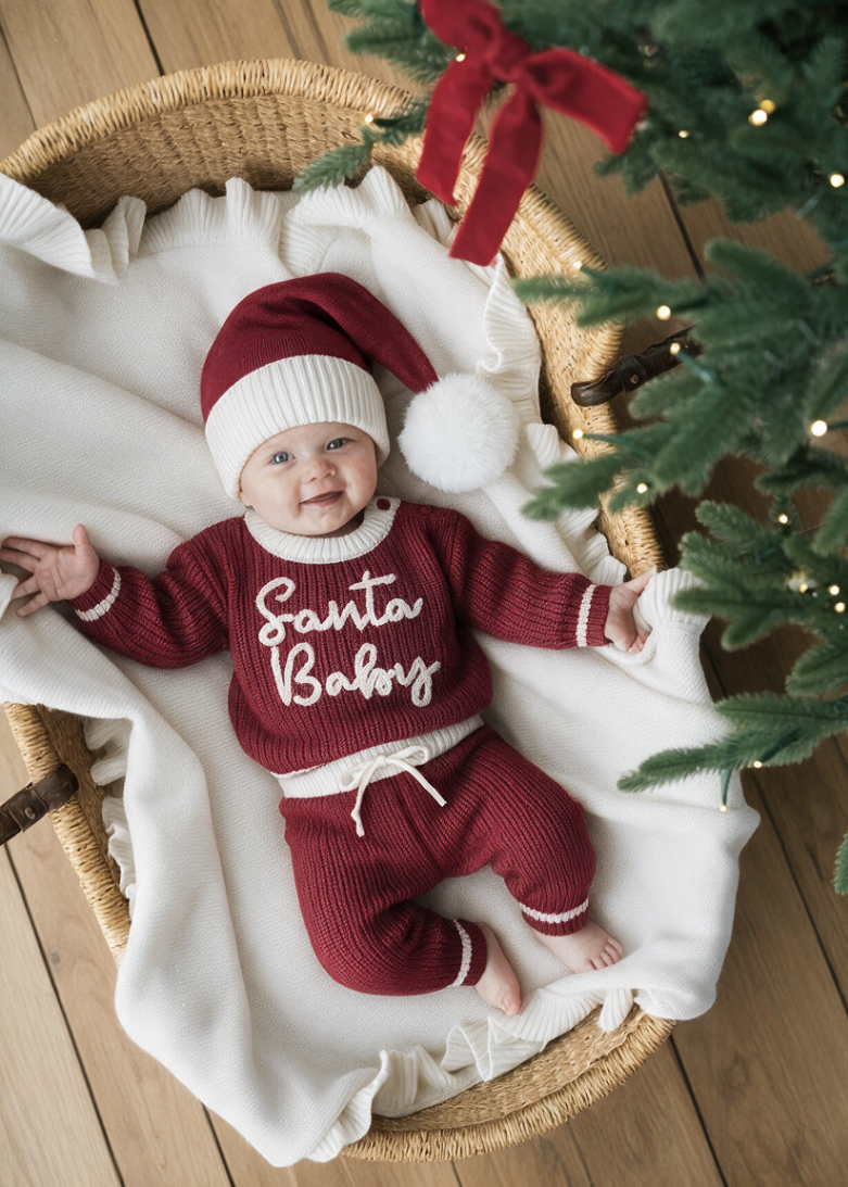 A baby wearing a maroon and white knit Santa Baby sweater and matching hat lies on a white blanket inside a woven basket next to a Christmas tree from Sweet June